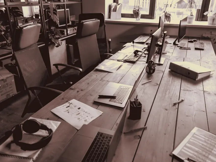 A long shared work table with monitors, notebooks, and empty chairs.